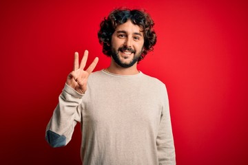 Young handsome man with beard wearing casual sweater standing over red background showing and pointing up with fingers number three while smiling confident and happy.