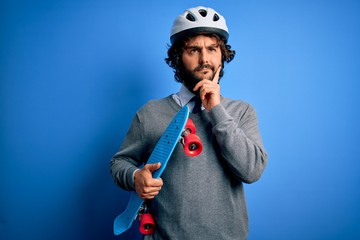 Handsome skater man with beard wearing security helmet holding skate over blue background serious face thinking about question, very confused idea