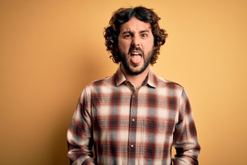 Young handsome man with beard wearing casual shirt standing over yellow background sticking tongue out happy with funny expression. Emotion concept.