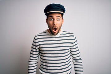 Young handsome sailor man with beard wearing navy striped uniform and captain hat afraid and shocked with surprise expression, fear and excited face. - obrazy, fototapety, plakaty