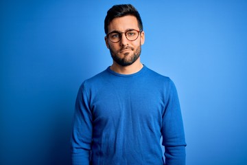 Young handsome man with beard wearing casual sweater and glasses over blue background looking...