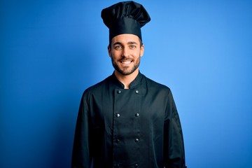 Young handsome chef man with beard wearing cooker uniform and hat over blue background with a happy and cool smile on face. Lucky person.