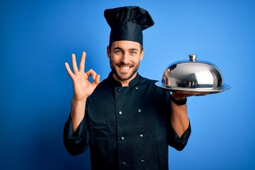 Young cooker man with beard wearing uniform holding tray with dome over blue background doing ok sign with fingers, excellent symbol