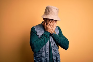 Handsome tourist man with beard on vacation wearing explorer hat over yellow background with sad...