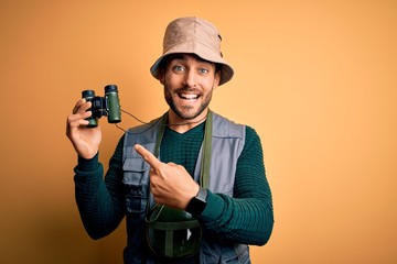 Young handsome tourist man with beard on vacation wearing explorer hat using binoculars very happy pointing with hand and finger