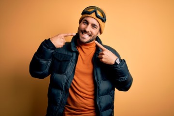 Young handsome skier man with beard wearing snow sportswear and ski goggles smiling cheerful showing and pointing with fingers teeth and mouth. Dental health concept.