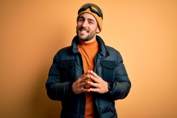 Young handsome skier man with beard wearing snow sportswear and ski goggles Hands together and fingers crossed smiling relaxed and cheerful. Success and optimistic