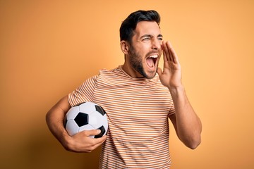 Fototapeta Handsome player man with beard playing soccer holding footballl ball over yellow background shouting and screaming loud to side with hand on mouth. Communication concept. obraz