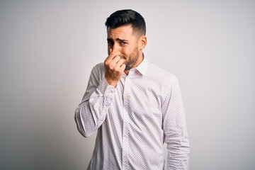 Young handsome man wearing elegant shirt standing over isolated white background smelling something stinky and disgusting, intolerable smell, holding breath with fingers on nose. Bad smell