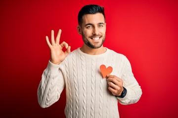 Young handsome man holding red shape heart as romantic and health symbol doing ok sign with fingers, excellent symbol