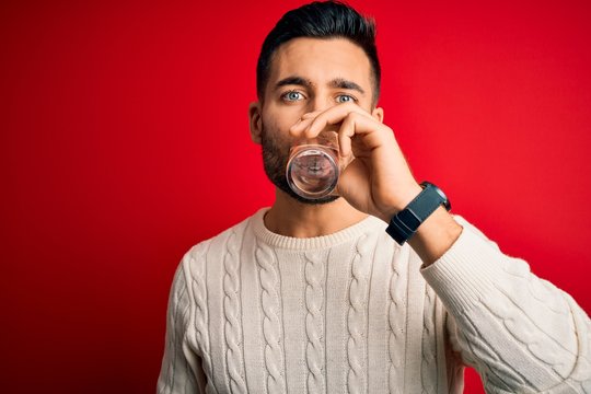 Young handsome man drinking glass of healthy water to refreshment standing over isolated red background