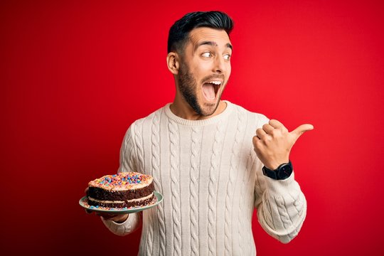 Young handsome man holding colorful birthday sweet cake over red isolated background pointing and showing with thumb up to the side with happy face smiling
