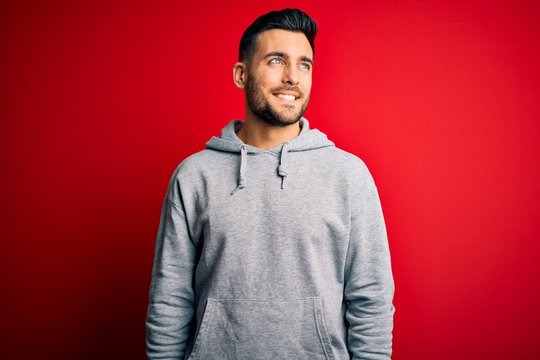 Young handsome sportsman wearing sweatshirt standing over isolated red background looking away to side with smile on face, natural expression. Laughing confident.