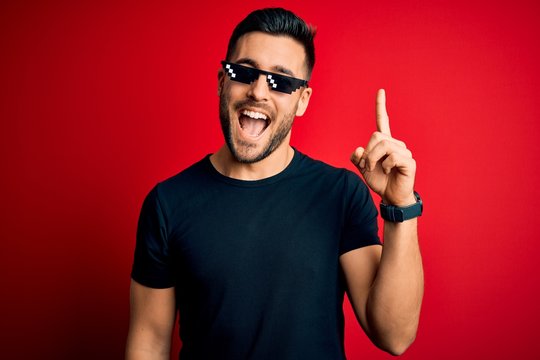 Young handsome man wearing funny thug life sunglasses over isolated red background pointing finger up with successful idea. Exited and happy. Number one.