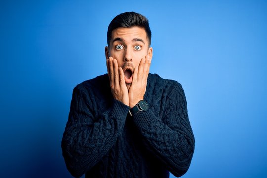Young handsome man wearing casual sweater standing over isolated blue background afraid and shocked, surprise and amazed expression with hands on face