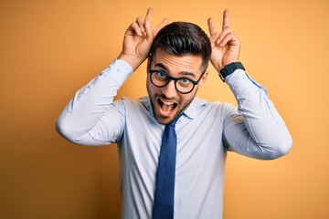 Young handsome businessman wearing tie and glasses standing over yellow background Posing funny and crazy with fingers on head as bunny ears, smiling cheerful