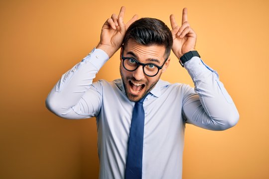 Young handsome businessman wearing tie and glasses standing over yellow background Posing funny and crazy with fingers on head as bunny ears, smiling cheerful
