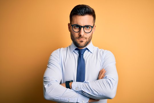 Young handsome businessman wearing tie and glasses standing over yellow background skeptic and nervous, disapproving expression on face with crossed arms. Negative person.