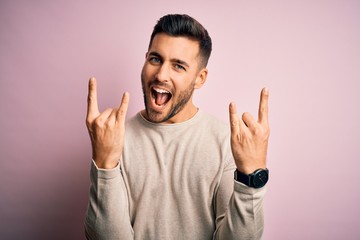 Young handsome man wearing casual sweater standing over isolated pink background shouting with crazy expression doing rock symbol with hands up. Music star. Heavy concept.