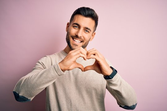 Young handsome man wearing casual sweater standing over isolated pink background smiling in love doing heart symbol shape with hands. Romantic concept.
