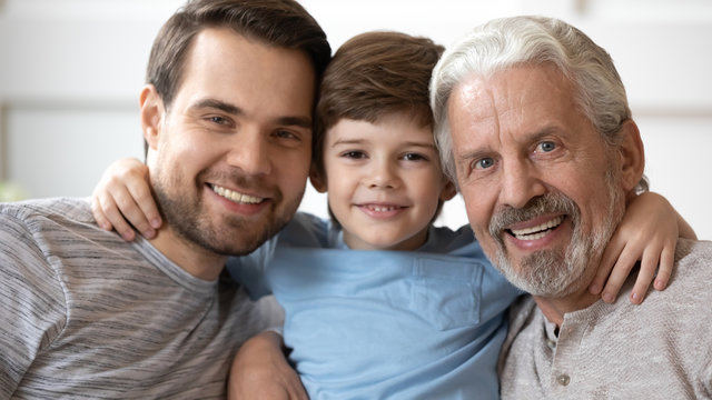 Headshot portrait of smiling three generations of men hug posing for picture together at home, happy little boy embrace young father and elderly grandfather look at camera relax in living room