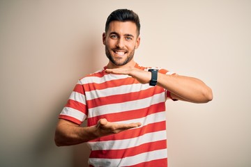 Young handsome man wearing casual striped t-shirt standing over isolated white background gesturing with hands showing big and large size sign, measure symbol. Smiling looking at the camera. Measuring
