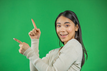 Portrait of beautiful 20s Asian woman wearing adorable dress expressing happiness pointing fingers sideways at copyspace isolated on green background