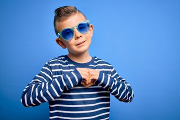 Young little caucasian kid with blue eyes standing wearing sunglasses over blue background smiling in love showing heart symbol and shape with hands. Romantic concept.