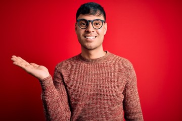 Young handsome hispanic man wearing nerd glasses over red background smiling cheerful presenting and pointing with palm of hand looking at the camera.
