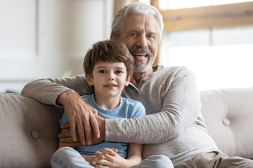 Portrait of smiling elderly grandfather relax on couch in living room hug little grandson, happy...
