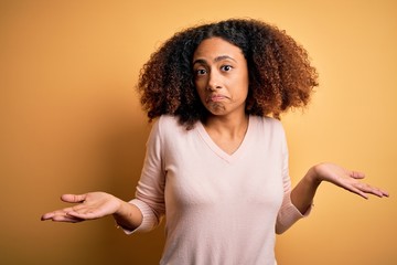 Young african american woman with afro hair wearing casual sweater over yellow background clueless and confused expression with arms and hands raised. Doubt concept.