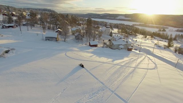 Aerial of a snowmobile driving trough fresh snow in a small village during golden hour 4K