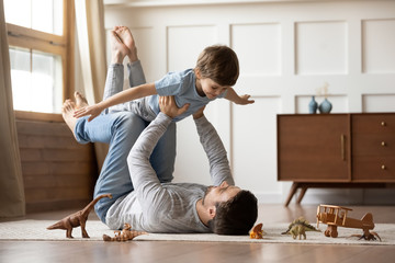 Happy young father lying on floor in living room hold fly with little preschooler son engaged in...