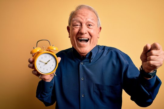 Senior grey haired man holding vintage alarm clock over yellow background screaming proud and celebrating victory and success very excited, cheering emotion