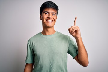 Fototapeta premium Young handsome man wearing casual t-shirt standing over isolated white background pointing finger up with successful idea. Exited and happy. Number one.