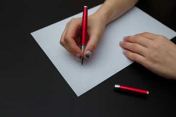 Female hands with fir-tree forest manicure holding red pen over white paper on black background. Christmas or winter nail art. Hands care, fingernails design, beauty and health concept. Copy space.