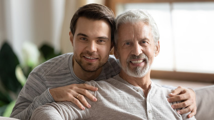 Portrait of smiling elderly father posing looking at camera hugging with adult grown-up son, happy young man embrace mature dad relax together at home on weekend, family value, relationships concept