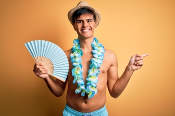 Young tourist man on vacation wearing swimwear and hawaiian lei flowers holding hand fan very happy...