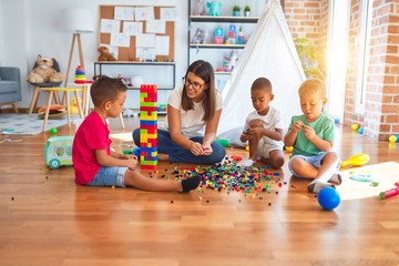 Young beautiful teacher and toddlers playing with building blocks around lots of toys at...