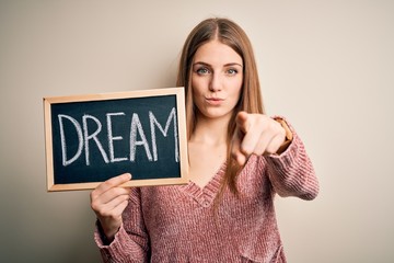 Young beautiful redhead woman holding blackboard with dream word message pointing with finger to the camera and to you, hand sign, positive and confident gesture from the front