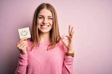 Young beautiful redhead woman holding reminder paper with smile emoji message doing ok sign with fingers, excellent symbol