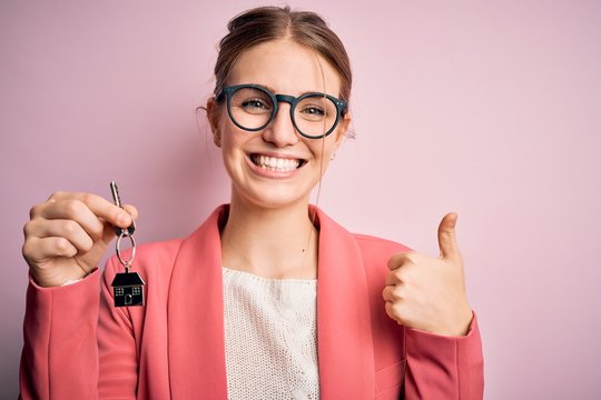 Young beautiful redhead house agent woman holding home key over pink bakcground happy with big smile doing ok sign, thumb up with fingers, excellent sign