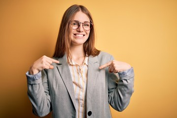 Fototapeta premium Young beautiful redhead woman wearing jacket and glasses over isolated yellow background looking confident with smile on face, pointing oneself with fingers proud and happy.