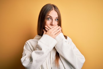 Young beautiful redhead sporty woman wearing sweatshirt over isolated yellow background shocked covering mouth with hands for mistake. Secret concept.