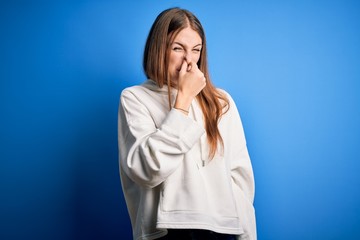 Young beautiful redhead sporty woman wearing sweatshirt over isolated blue background smelling something stinky and disgusting, intolerable smell, holding breath with fingers on nose. Bad smell