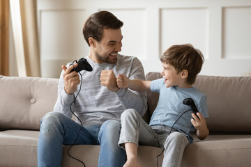 Overjoyed young father and little son sit relax on couch in living room playing computer games...