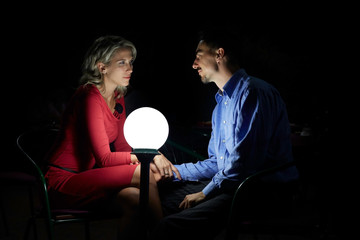 Beautiful blonde woman in red dress, and a man in blue shirt sits together in darkness, next to a ball shaped lamp. Couple dating at evening time.