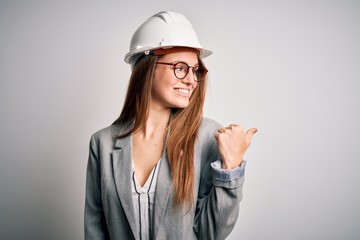 Young beautiful redhead architect woman wearing security helmet over white background smiling with happy face looking and pointing to the side with thumb up.