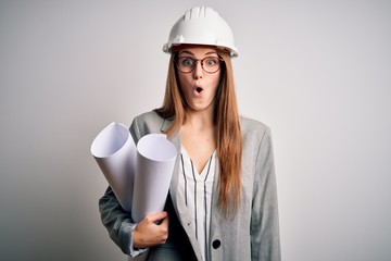 Young beautiful redhead architect woman wearing glasses and safety helmet holding blueprints scared in shock with a surprise face, afraid and excited with fear expression