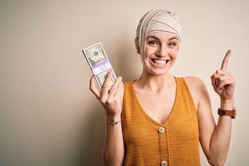 Young woman injured wearing bandage on head holding money from medical insurance payment surprised with an idea or question pointing finger with happy face, number one